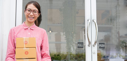 An Asian teenage girl with braces stands in front of glass doors holding a stack of boxes