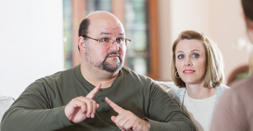 A man signs with a person sitting across from him while a woman looks over his shoulder