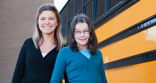 A white middle school girl stands with a white woman in front of a school bus.