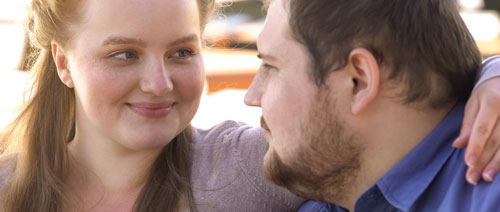 A white woman with long reddish hair smiles as she puts her arm around the shoulders of a white man with brown hair and a beard